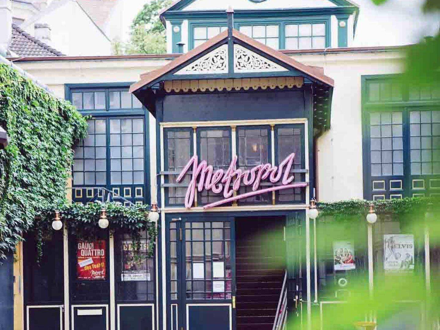 Historische Gebäudefassade mit einem leuchtenden 'Metropol'-Neonschild, umgeben von üppigem Grün und dekorativen Elementen.