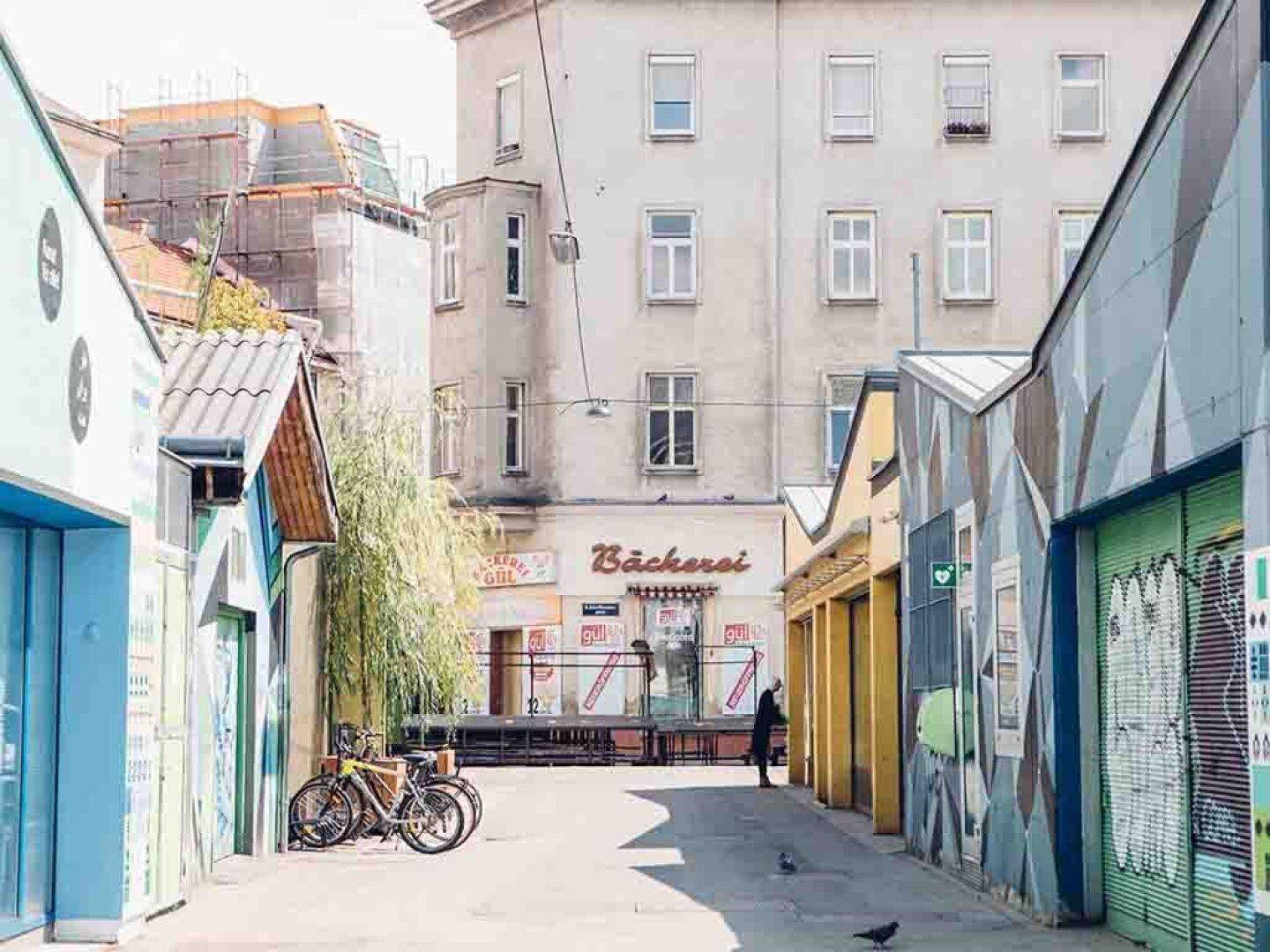 Belebte Gasse mit verschiedenen Gebäuden, einer Bäckerei und Fahrrädern, die eine urbane Atmosphäre schaffen.