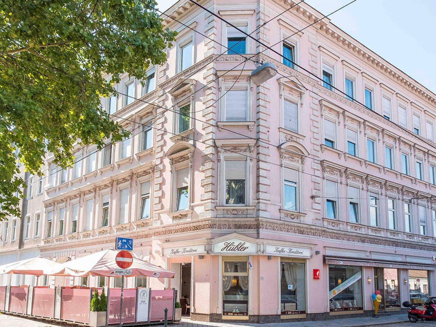 Historisches Eckgebäude in Rosa mit vielen Fenstern und einem Straßencafé im Erdgeschoss in einer urbanen Umgebung.