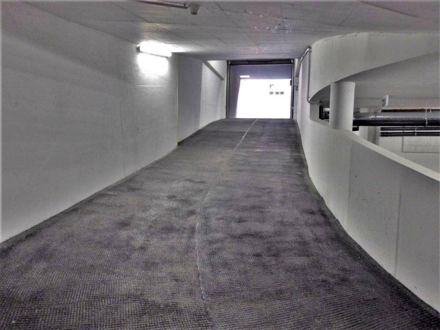 Zufahrtsrampe zu einer Tiefgarage mit offenem Tor am Ende, beleuchtet von Wandlampen und Tageslicht.