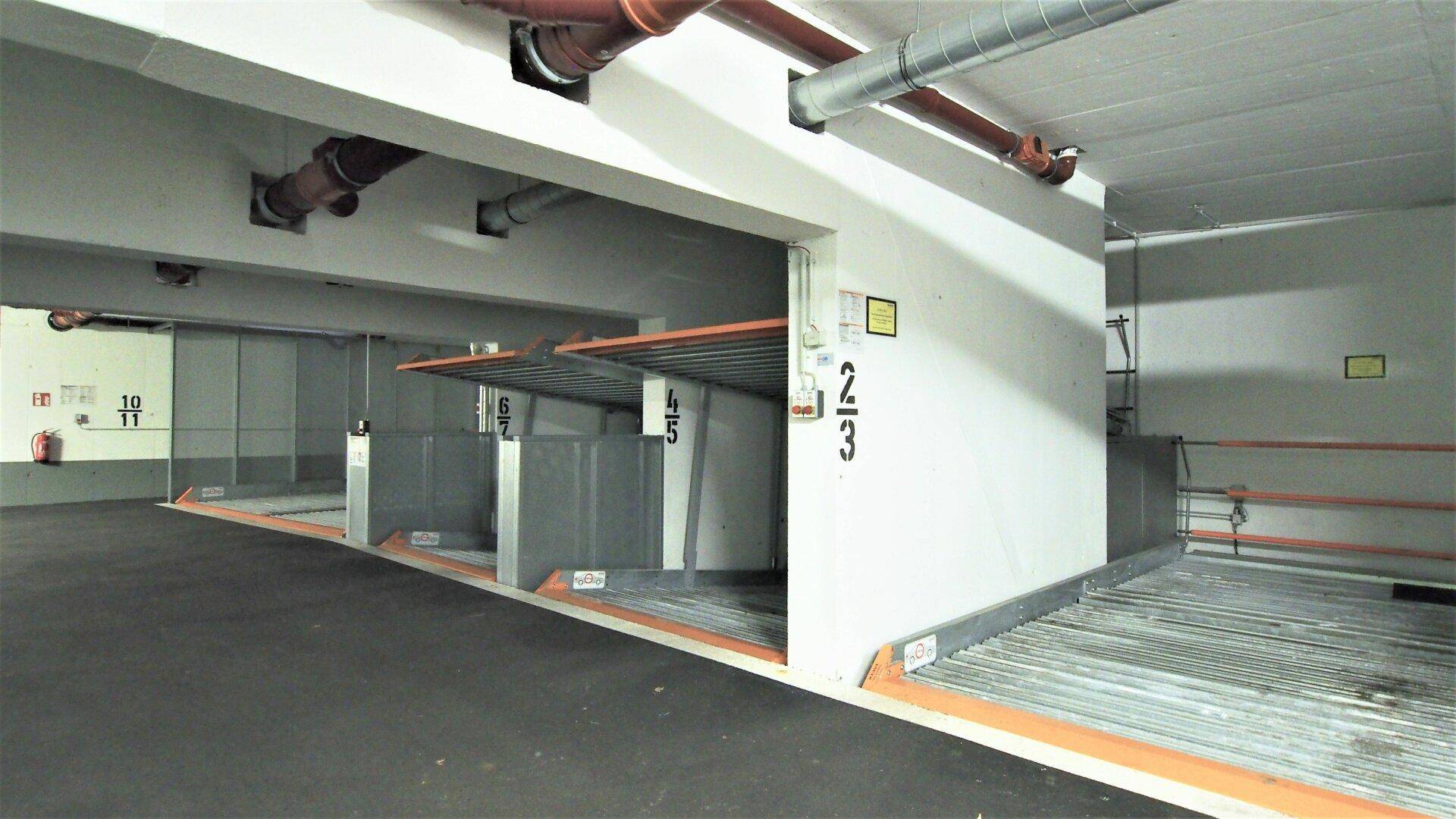 Tiefgarage mit nummerierten mechanischen Parkplätzen und sichtbaren Installationen an der Decke.