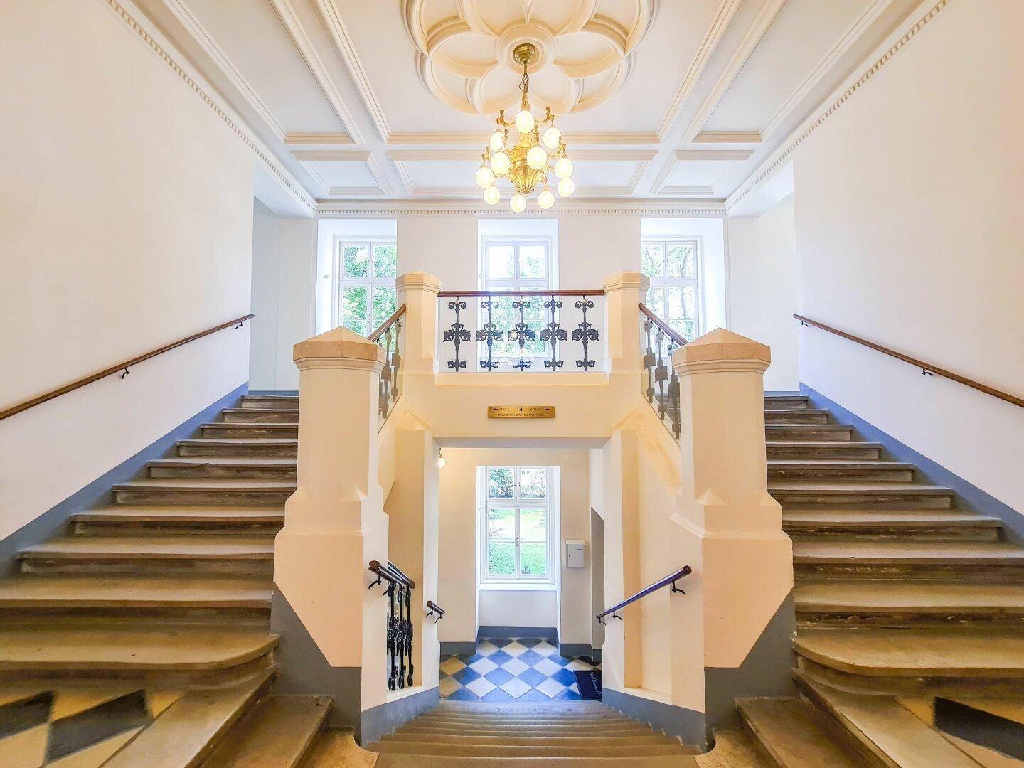 Eindrucksvolles Treppenhaus mit klassischer Architektur, hohen Decken, Kronleuchter und großen Fenstern, die viel natürliches Licht hereinlassen.