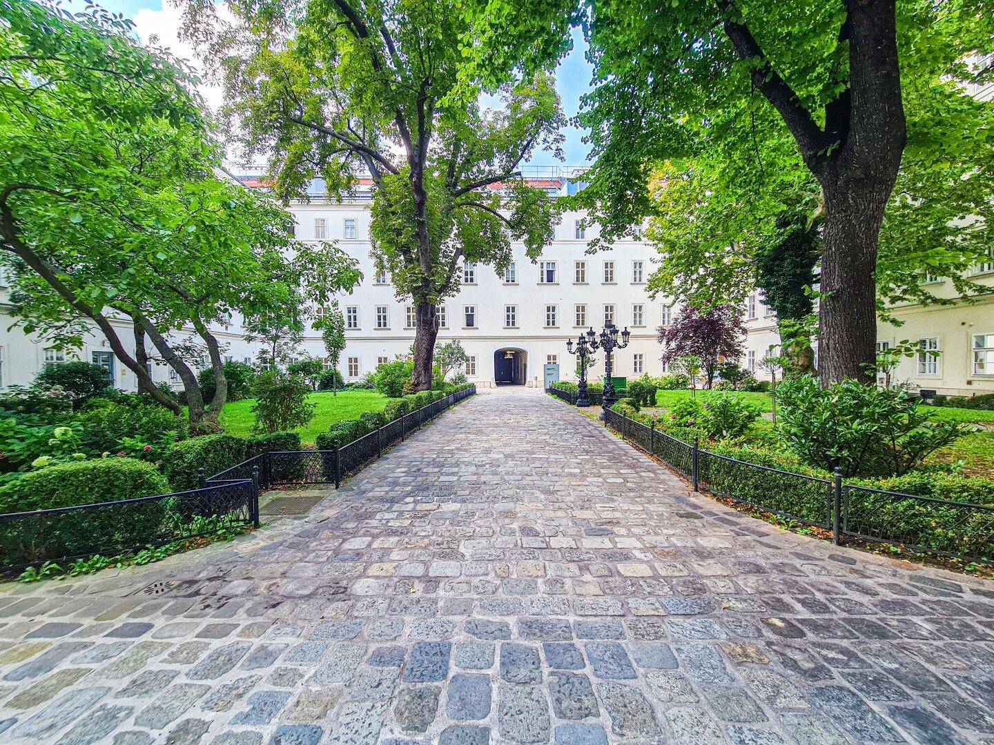 Ein Kopfsteinpflasterweg führt durch einen gepflegten grünen Innenhof zu einem eleganten weißen Gebäude mit vielen Fenstern.