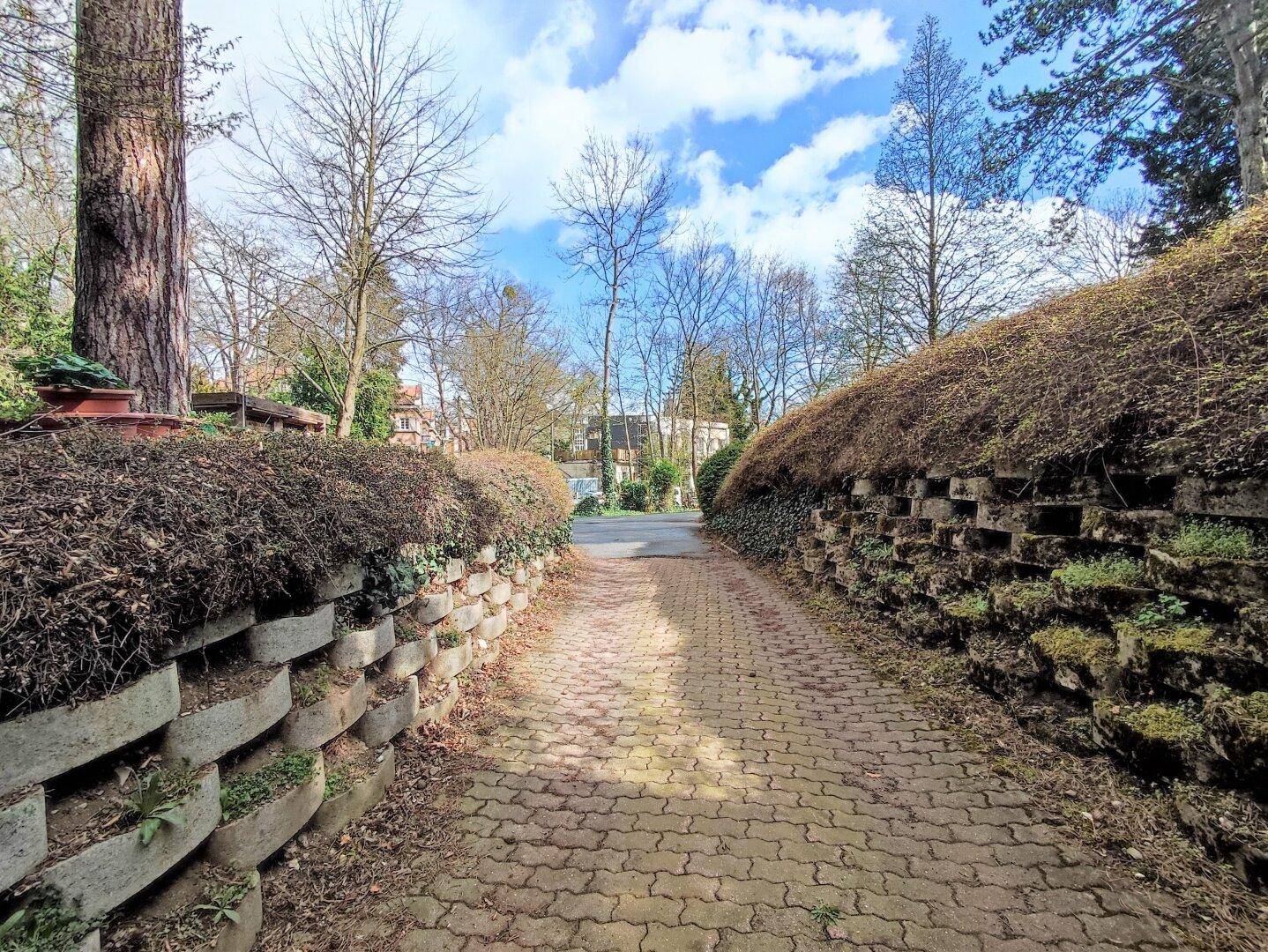 Gepflasterte Auffahrt umgeben von begrünten Stützmauern und Bäumen unter blauem Himmel.