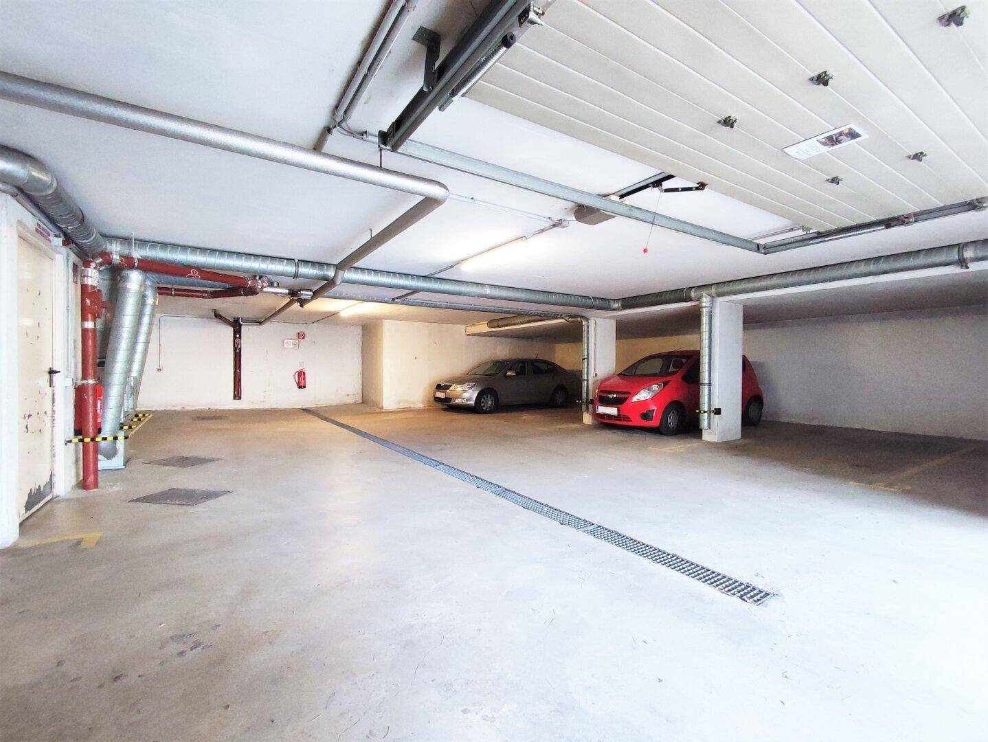 Geräumige Tiefgarage mit zwei geparkten Autos und sichtbaren Versorgungsleitungen an der Decke.