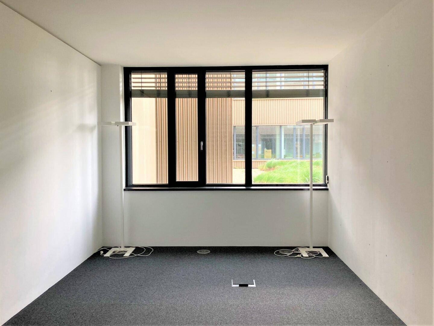 Kompakter Büroraum mit Teppichboden und einem großen Fenster, das den Blick auf ein anderes Gebäude freigibt.
