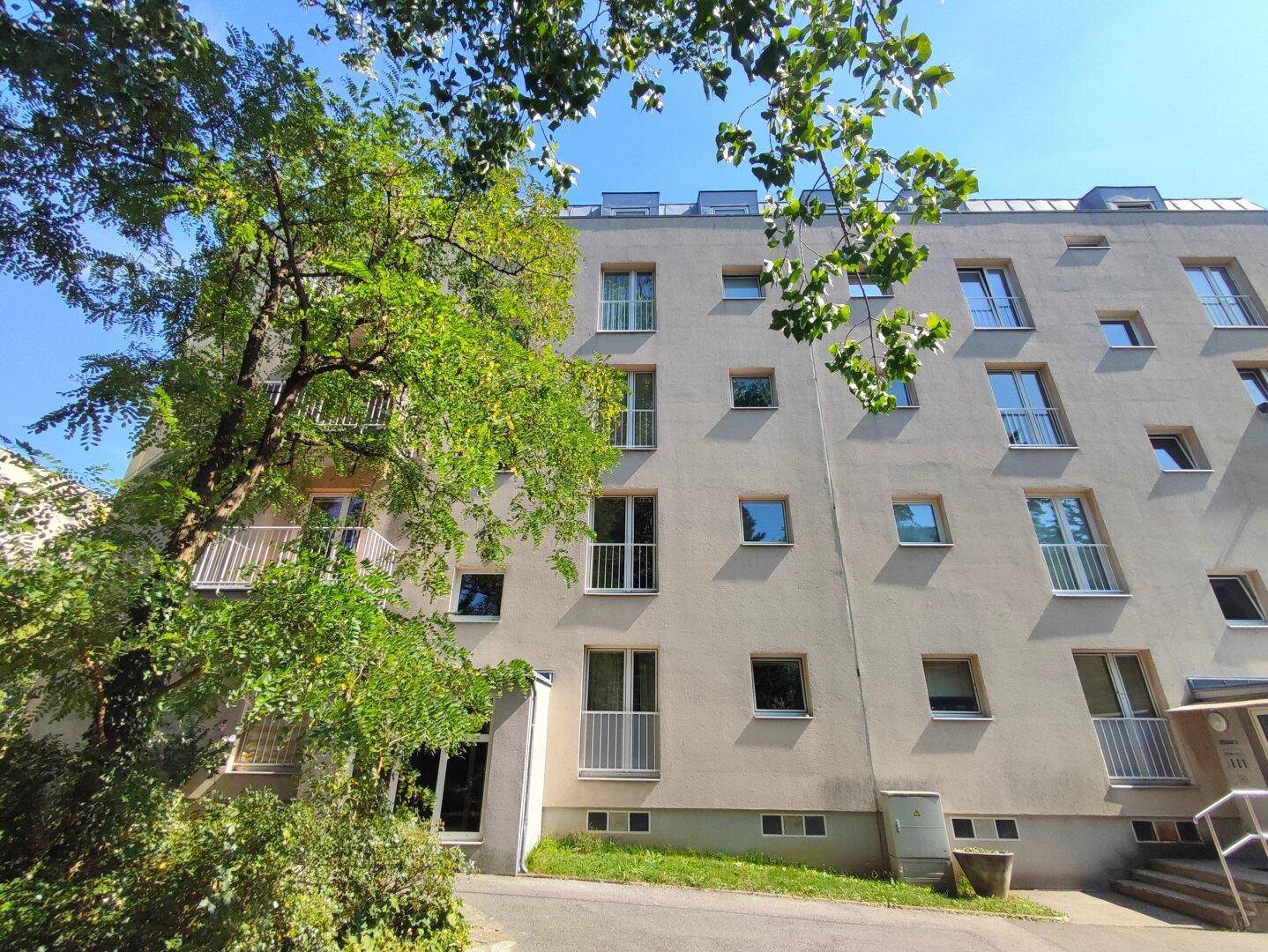 Fassade eines Mehrfamilienhauses mit vielen Fenstern und Balkonen, umgeben von Bäumen.