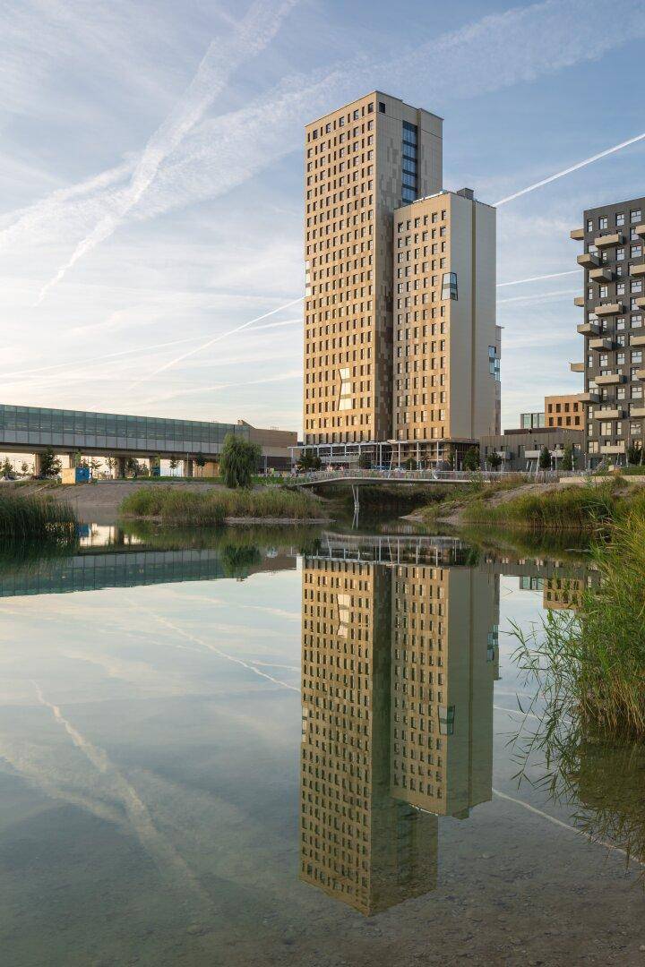 Hohes Gebäude spiegelt sich in einem ruhigen Gewässer wider, umgeben von Natur und Brücken.