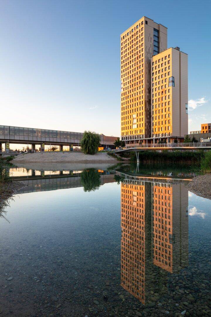 Hohes Gebäude spiegelt sich im ruhigen Wasser, umgeben von einer Brücke und grüner Ufervegetation.