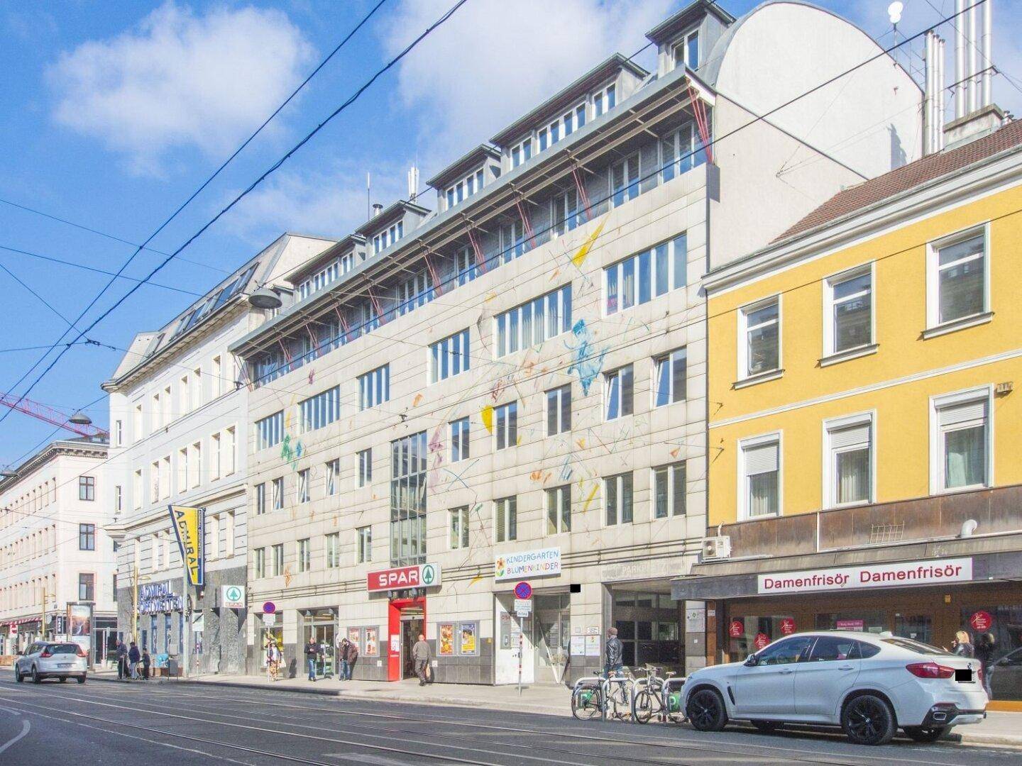 Mehrstöckiges Gebäude mit moderner Fassade, Geschäften im Erdgeschoss und einer belebten Straße davor unter blauem Himmel.