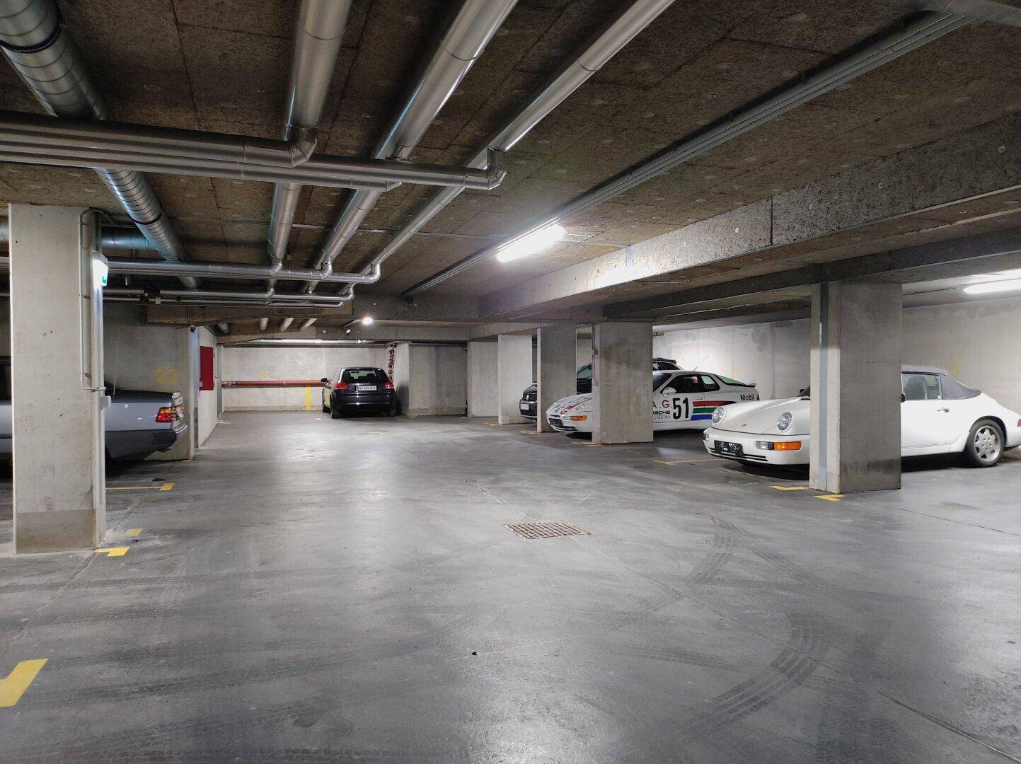Weitläufige Tiefgarage mit Betonboden und Decke, beleuchtet von Leuchtstoffröhren.