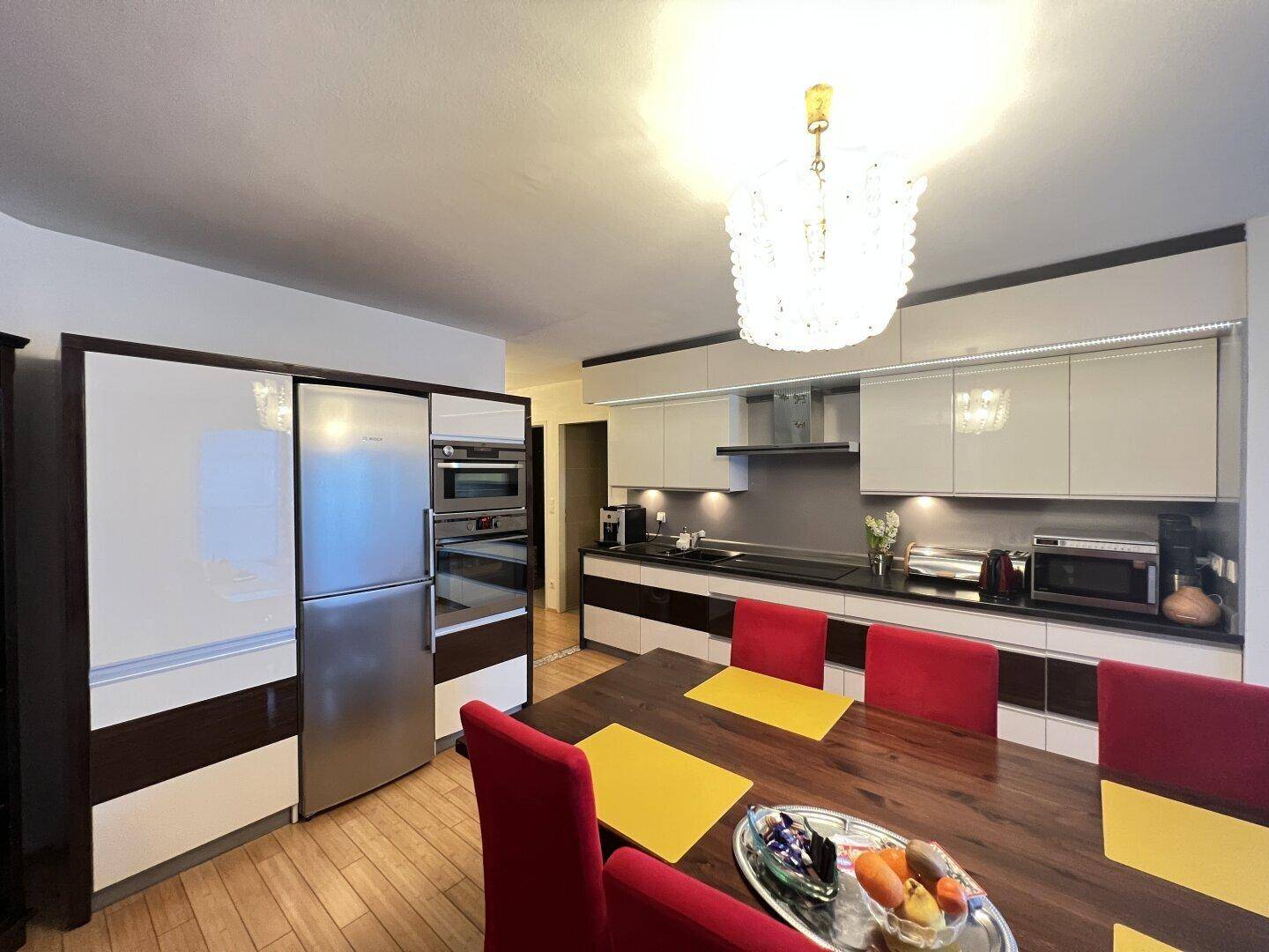 Essbereich mit Holztisch und roter Bestuhlung, angrenzend an die moderne Küche mit Geräten.