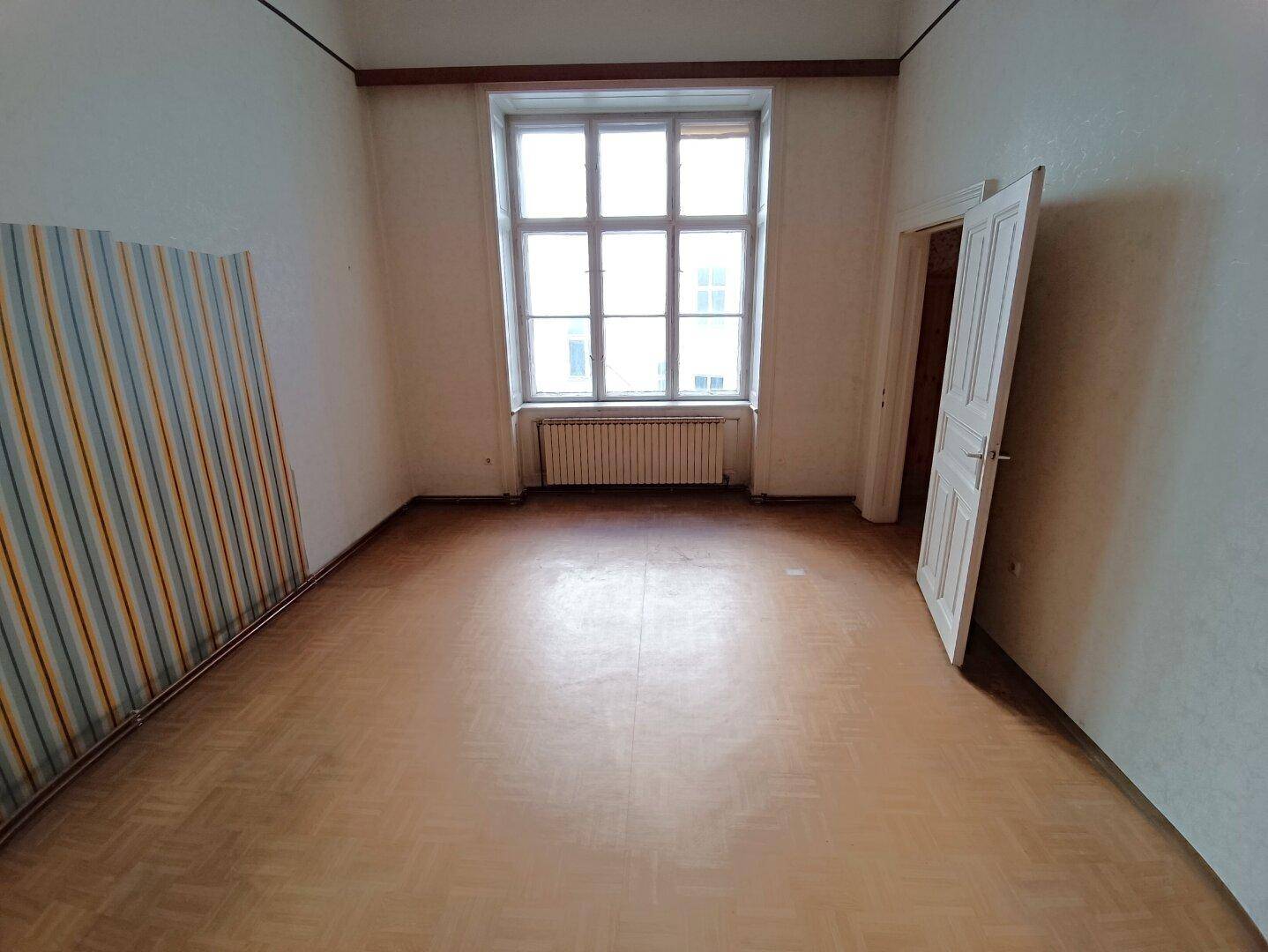Leerer Raum mit großem Fenster, Parkettboden und gestreifter Tapete an einer Wand.