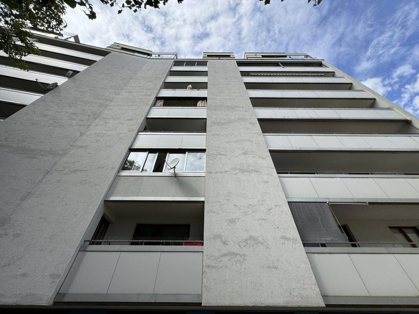 Hohes Wohngebäude mit grauer und weißer Fassade sowie zahlreichen Balkonen unter bewölktem Himmel.