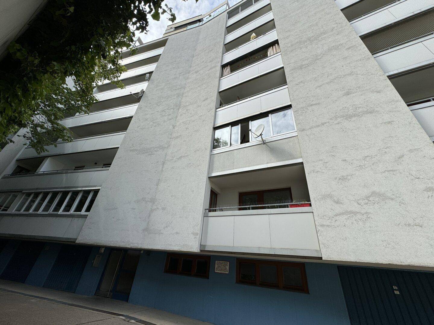 Ansicht eines mehrstöckigen Wohnhauses mit grauer Fassade und Balkonen, umgeben von Bäumen.