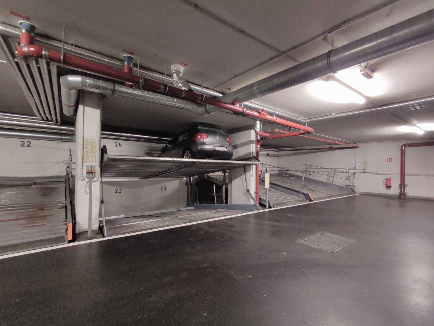 Tiefgarage mit einem auf einer oberen Plattform geparkten Auto und sichtbaren Versorgungsleitungen an der Decke.