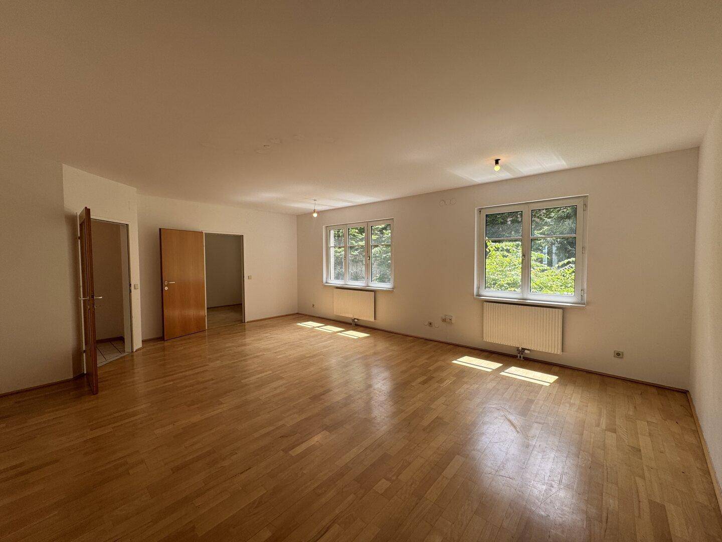 Helles Wohnzimmer mit Holzboden und zwei großen Fenstern, die einen Blick ins Grüne bieten.