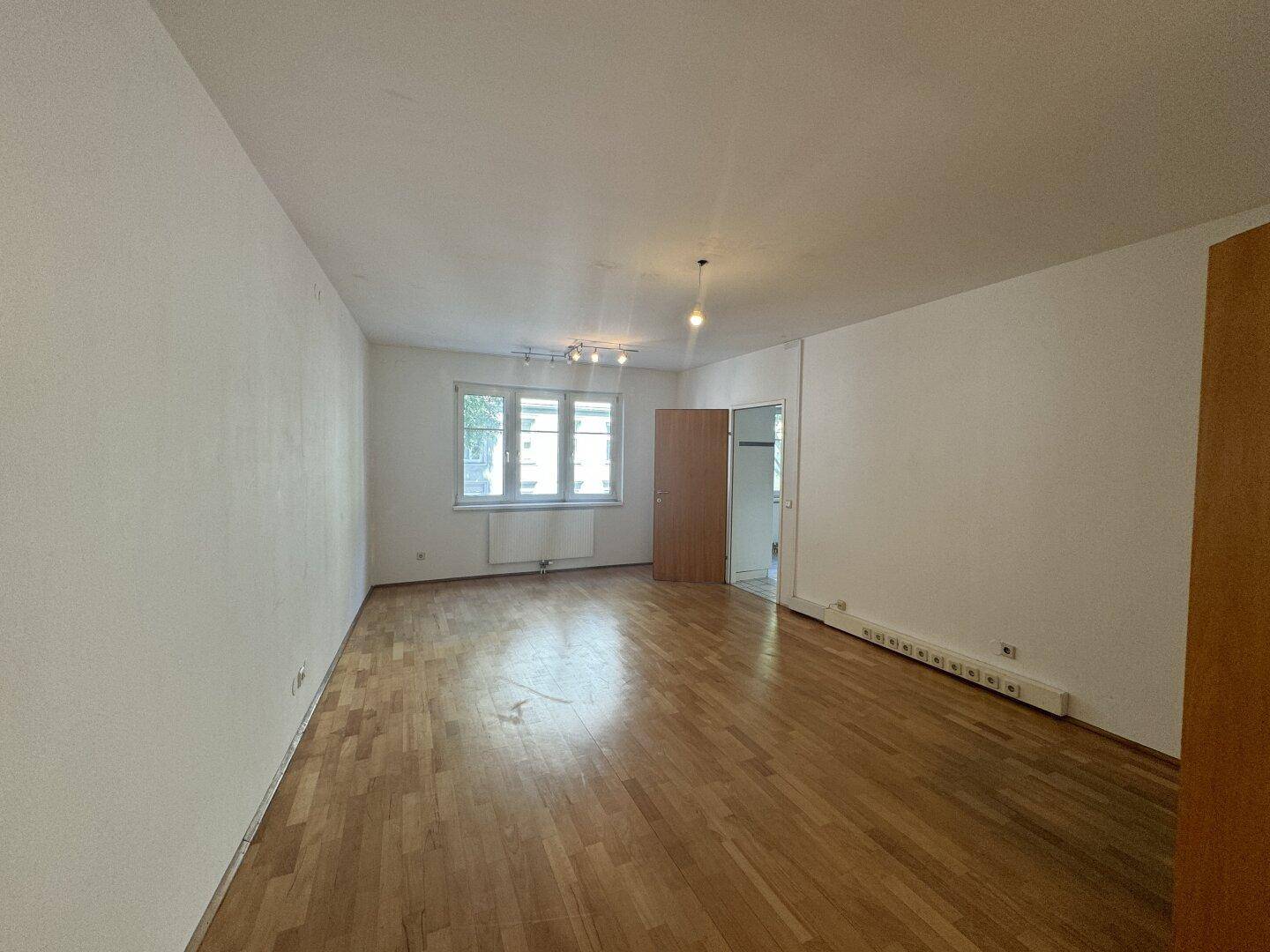 Geräumiges, leeres Wohnzimmer mit hellem Parkettboden und großen Fenstern, die viel Tageslicht hereinlassen.