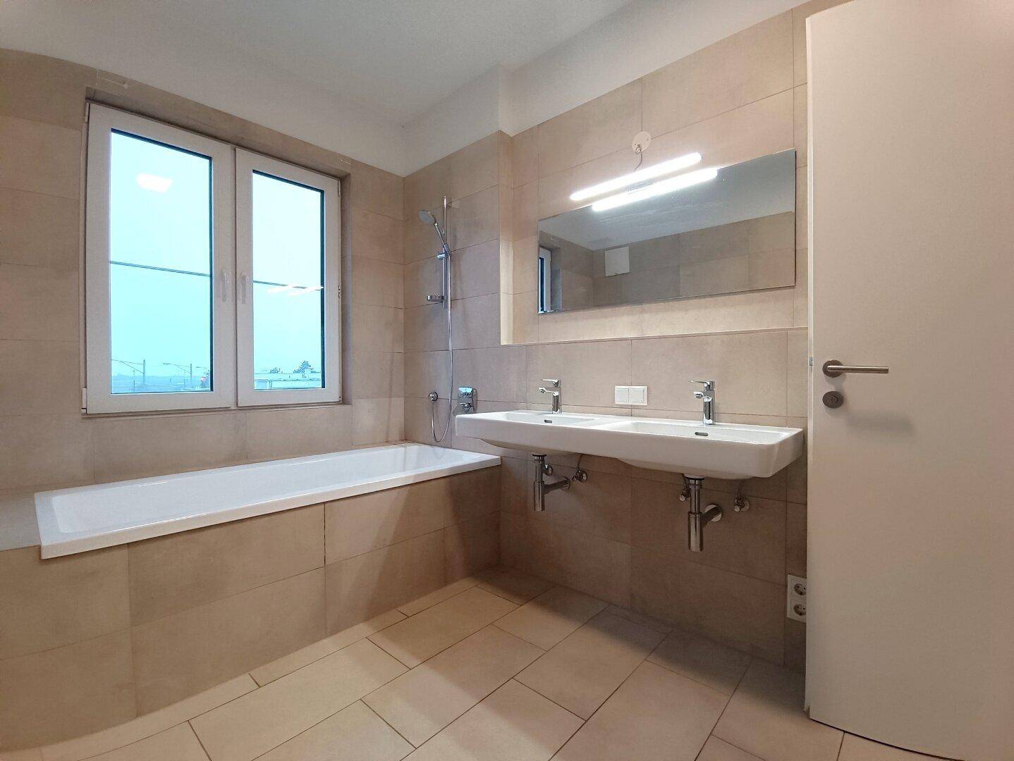 Modernes Badezimmer mit beigen Fliesen, Badewanne, Doppelwaschbecken und Fenster.
