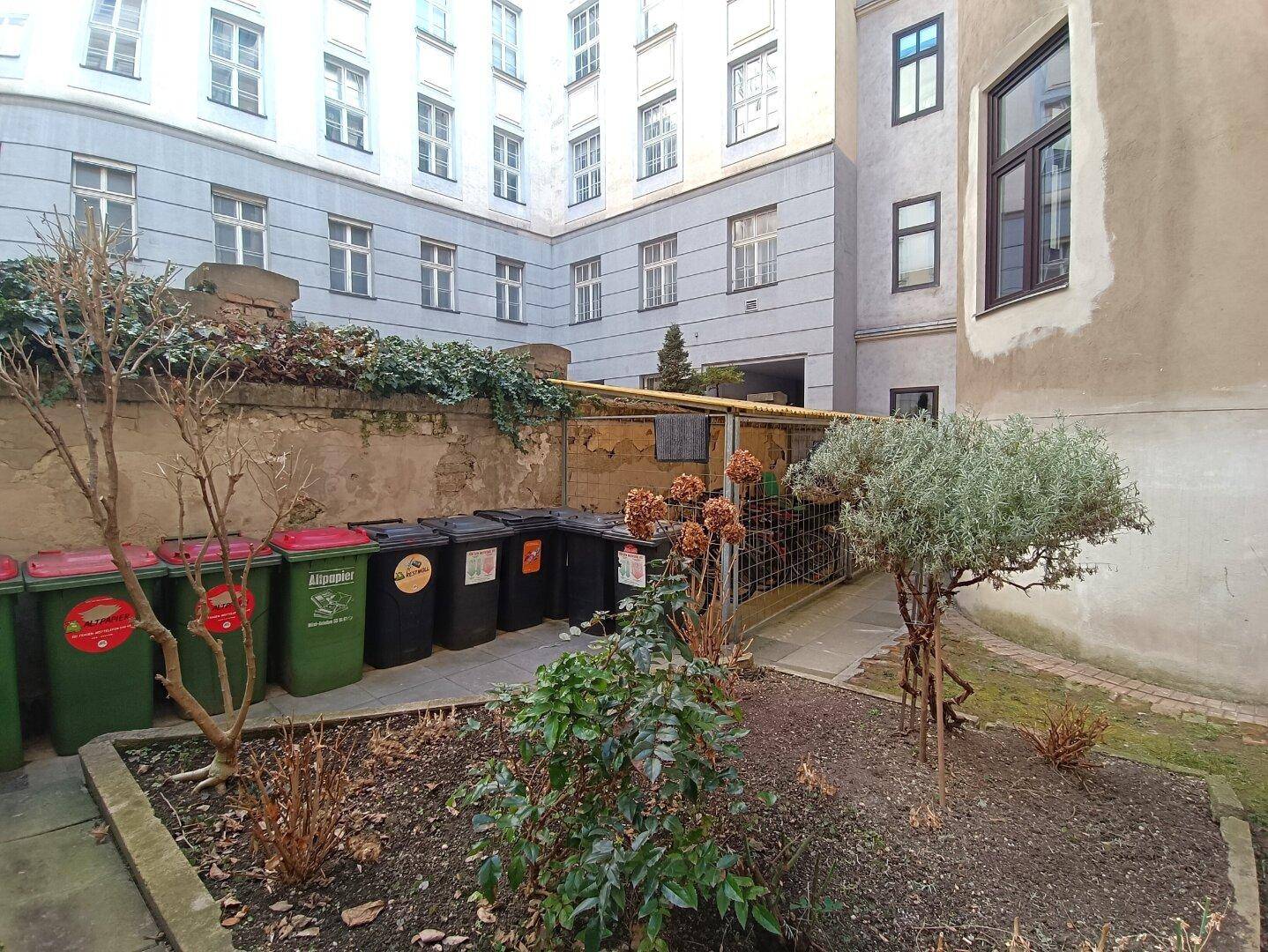 Ein weiterer Bereich des Innenhofs mit Mülltonnen und einigen Pflanzen, umgeben von Gebäudefassaden.