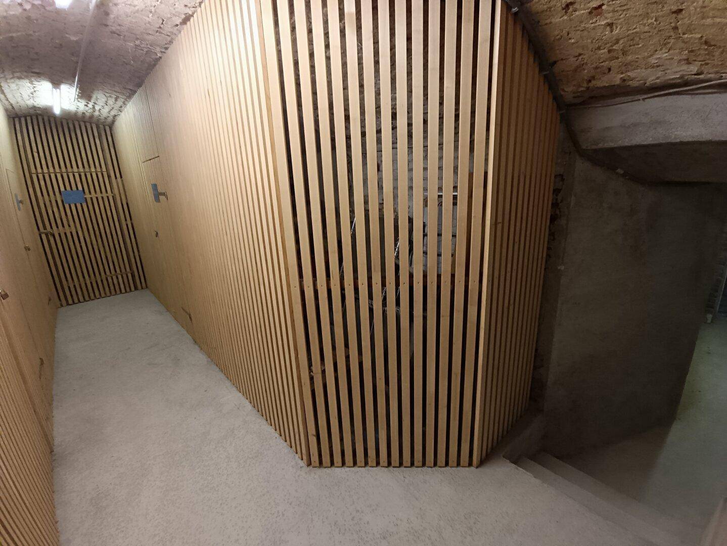 Ein modernisierter Kellergang mit hellen Holzlamellenwänden und Betonboden, der zu den Lagerräumen führt.