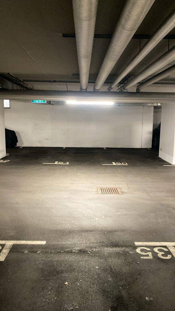 Leerer Tiefgaragenstellplatz mit sichtbaren Rohren an der Decke und heller Beleuchtung.