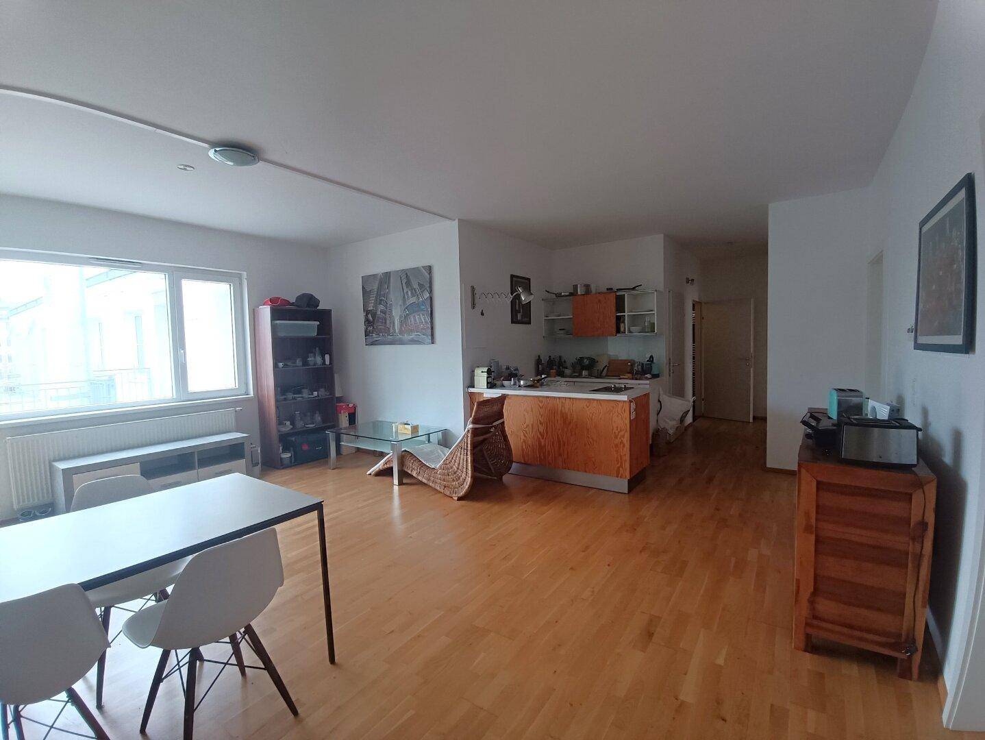 Geräumiger Wohnbereich mit großen Fenstern, Holzboden und offener Küchenzeile.