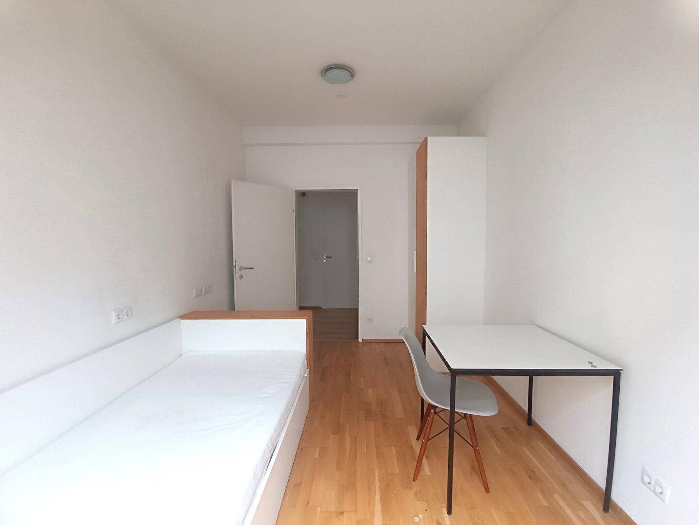 Einfaches Schlafzimmer mit Holzboden, Schreibtisch und Kleiderschrank.