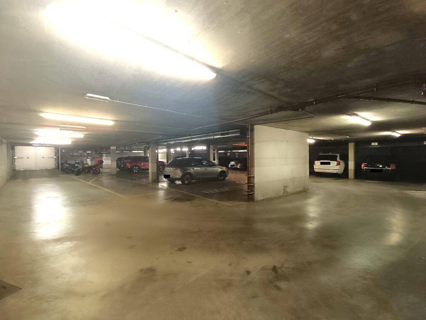 Weitläufige Tiefgarage mit mehreren geparkten Fahrzeugen und klarer Beleuchtung durch Leuchtstoffröhren.