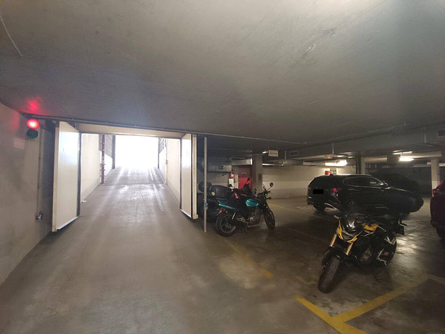 Einfahrt zur Tiefgarage mit geöffneten Toren und Motorrädern im Vordergrund, helles Licht am Ausgang.