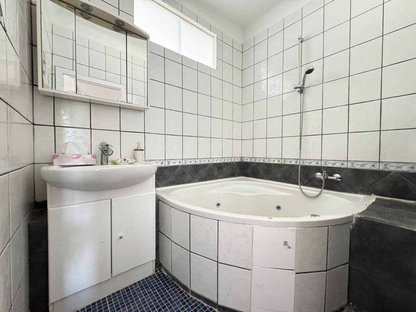 Badezimmer mit Eckbadewanne, Dusche, Waschbecken und Fliesen in Weiß und Dunkelgrau.