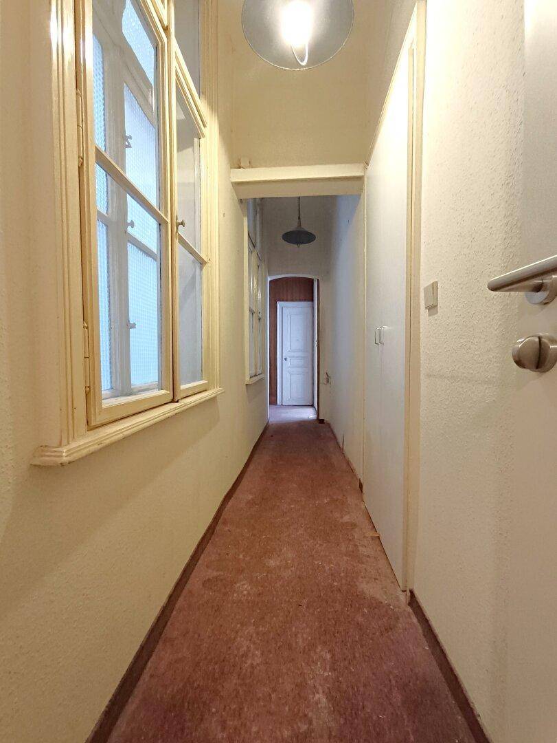 Langer, schmaler Flur mit Fenster und einfacher Beleuchtung, der renovierungsbedürftig ist.