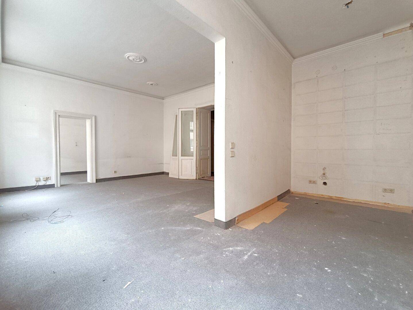 Großer Wohnbereich mit Teppichboden und hohen Decken, der auf eine Modernisierung wartet.