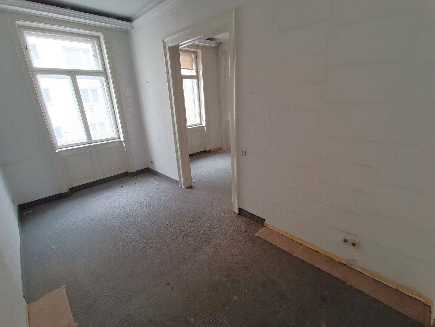 Heller Wohnraum im Rohzustand mit zwei großen Fenstern und hohen Decken.