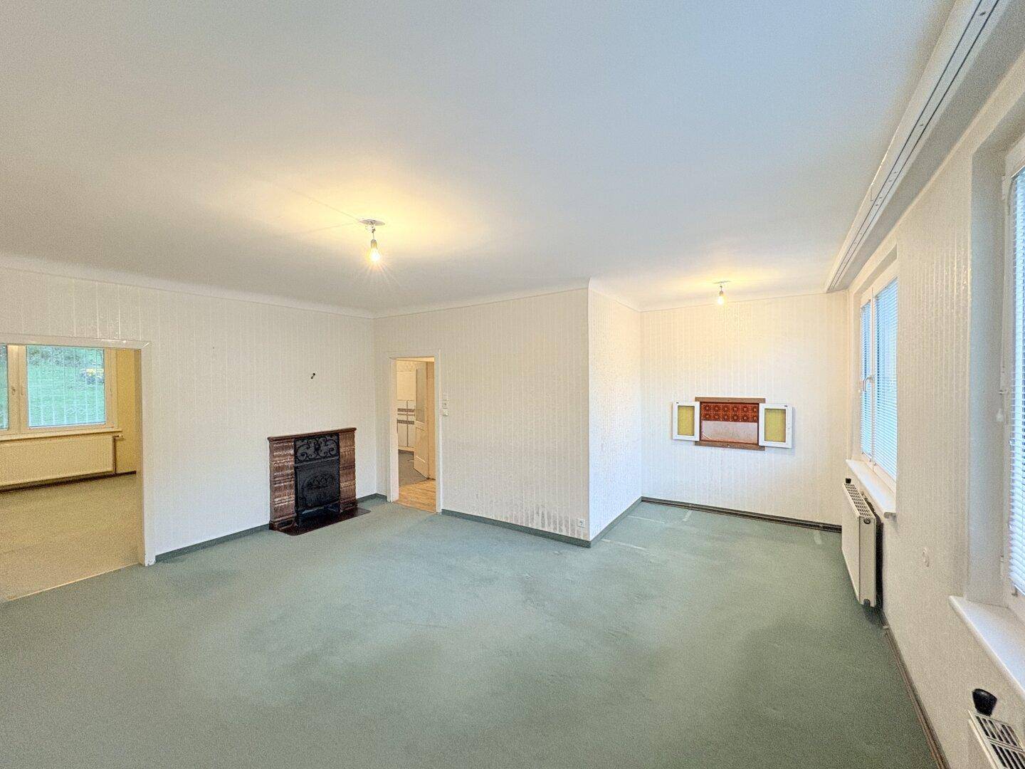 Geräumiges Wohnzimmer mit Kamin, grünem Teppichboden und Blick in einen weiteren Raum.