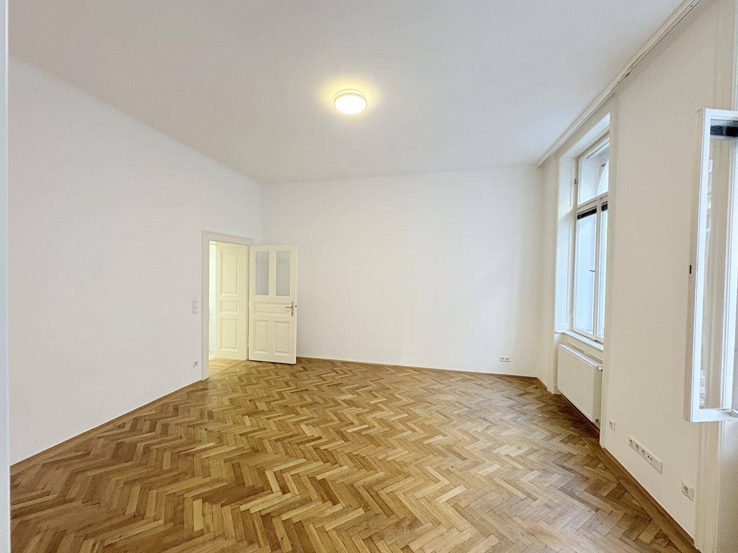 Großes, helles Zimmer mit klassischem Fischgrätparkett und hohen Decken.