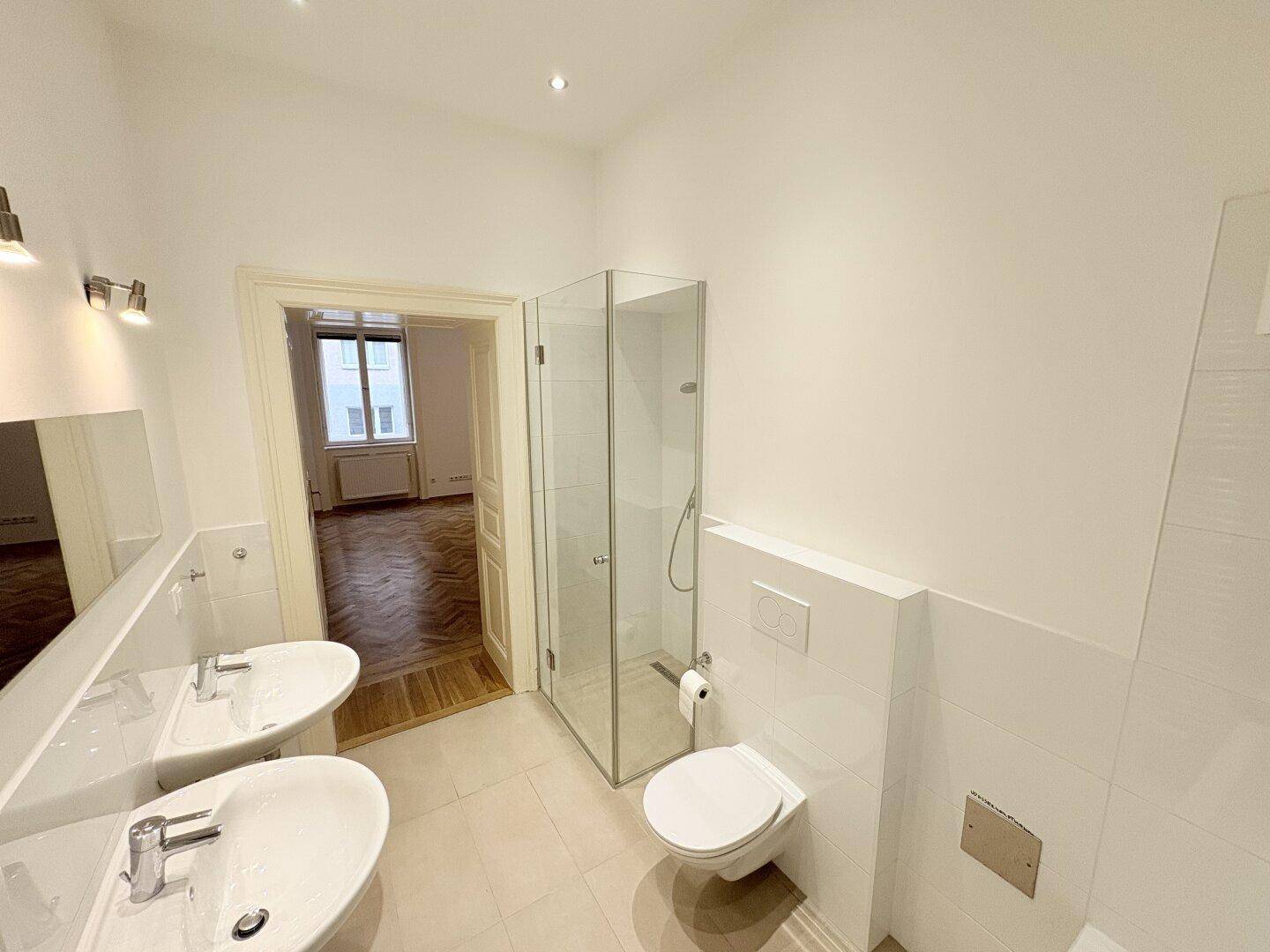 Helles Badezimmer mit Doppelwaschbecken, Duschkabine und Zugang zu einem weiteren Raum.