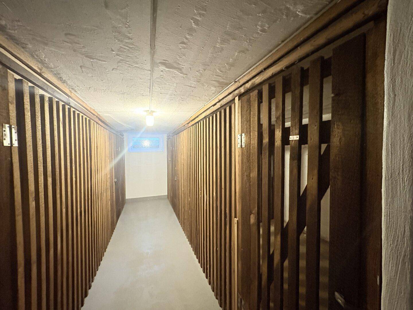 Langer Kellerflur mit beidseitigen Holzverschlägen für Lagerzwecke und einem kleinen Fenster am Ende.