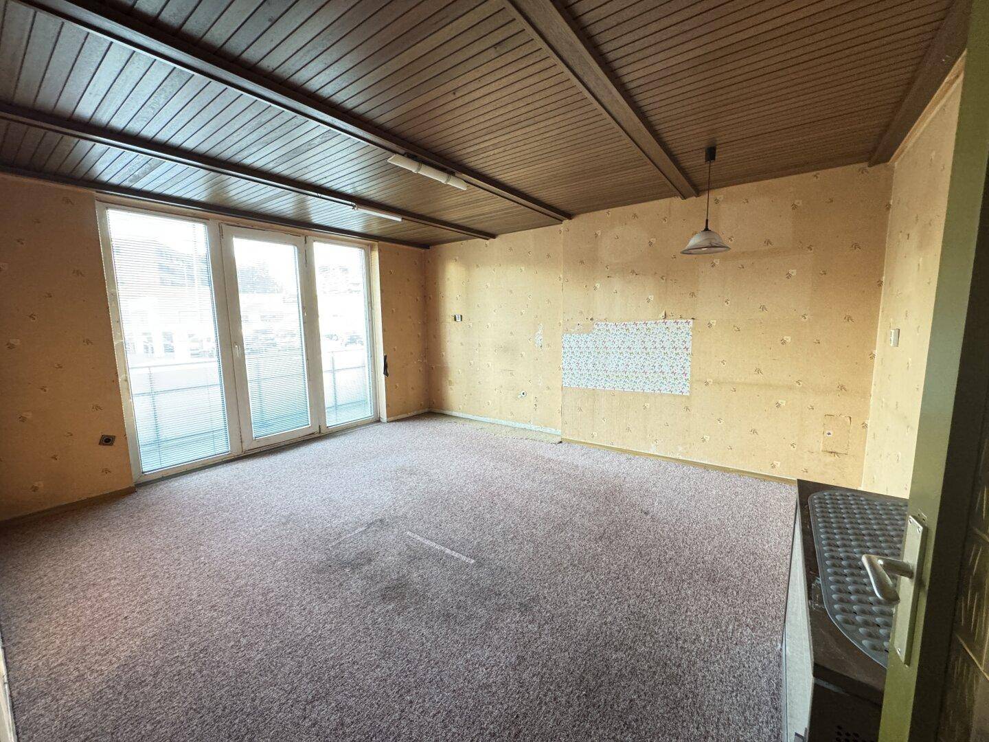 Leeres Wohnzimmer mit Teppichboden, Holzdecke und großen Fenstertüren zum Balkon.
