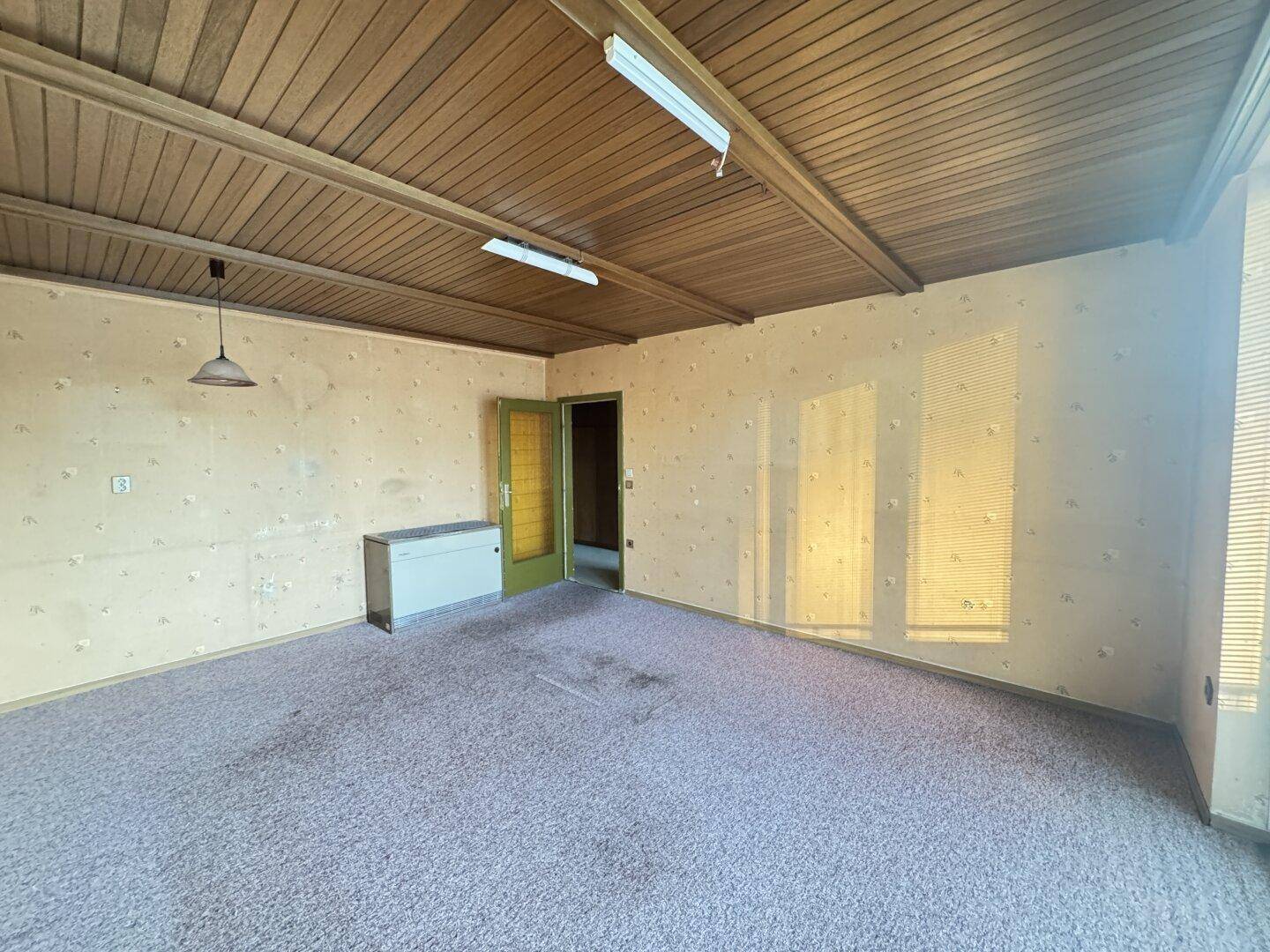 Geräumiges Wohnzimmer mit Teppichboden, Holzdecke und älterer Tapete, bereit zur Renovierung.