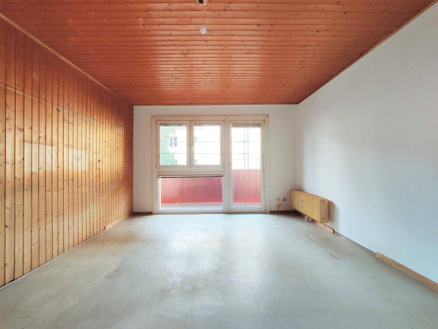 Leeres Wohnzimmer mit Holzdecke, Holzpaneelen an einer Wand und Zugang zu einem Balkon.