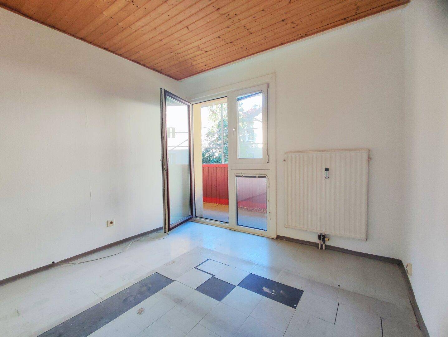 Leeres Schlafzimmer mit Holzdecke, Heizkörper und Zugang zu einem Balkon.