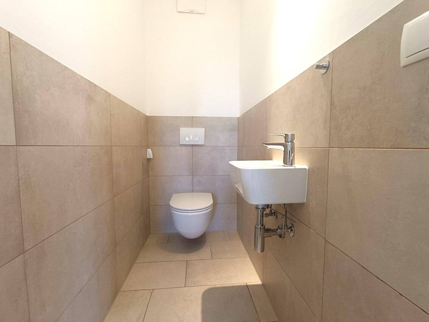 Modernes Gäste-WC mit Wand-WC, kleinem Waschbecken und beigefarbenen Fliesen.