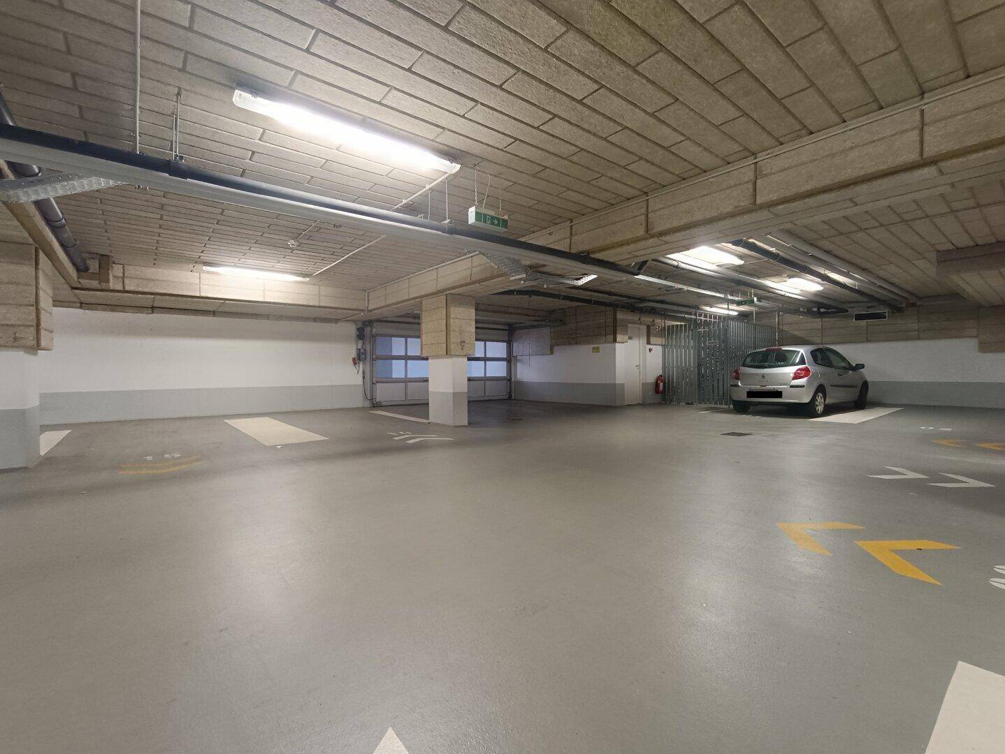 Helle Tiefgarage mit breiten Fahrwegen, markierten Stellplätzen und einem sichtbaren Zugangsbereich für Fahrzeuge.