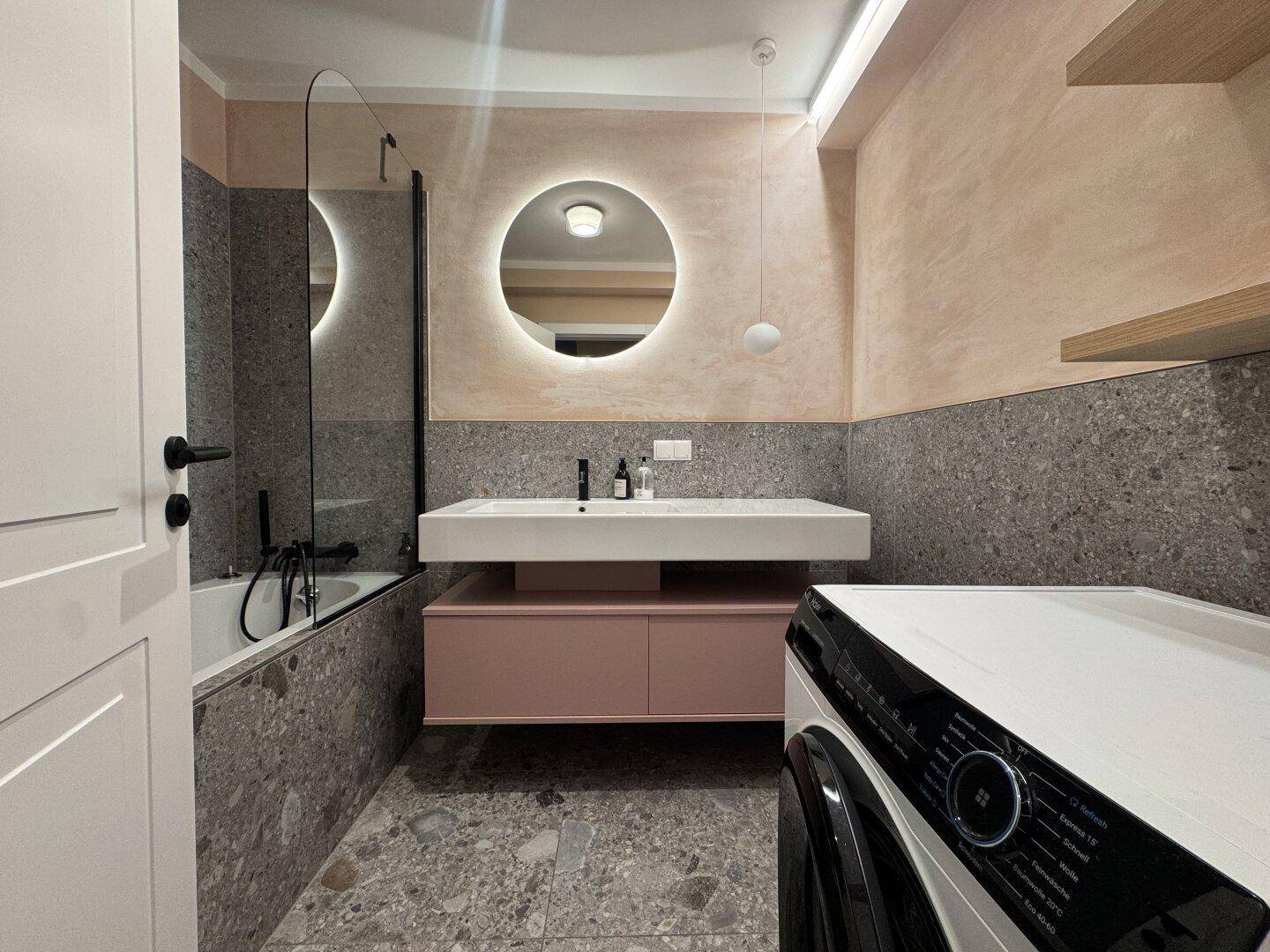 Modernes Badezimmer mit Terrazzo-Fliesen, beleuchtetem Spiegel, Badewanne und Waschmaschine.