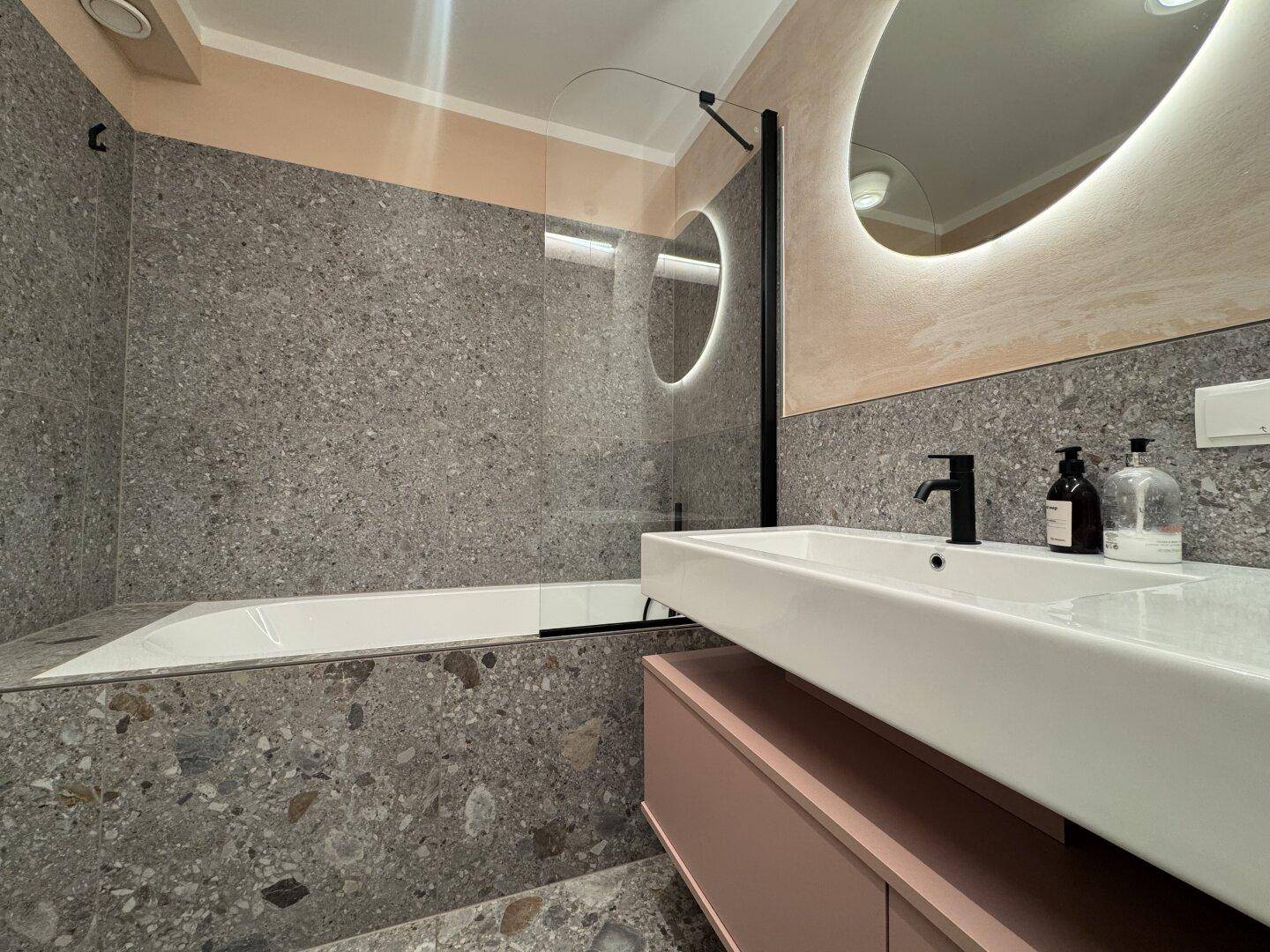 Modernes Badezimmer mit Terrazzo-Fliesen, Badewanne mit Duschabtrennung und beleuchtetem Spiegel.