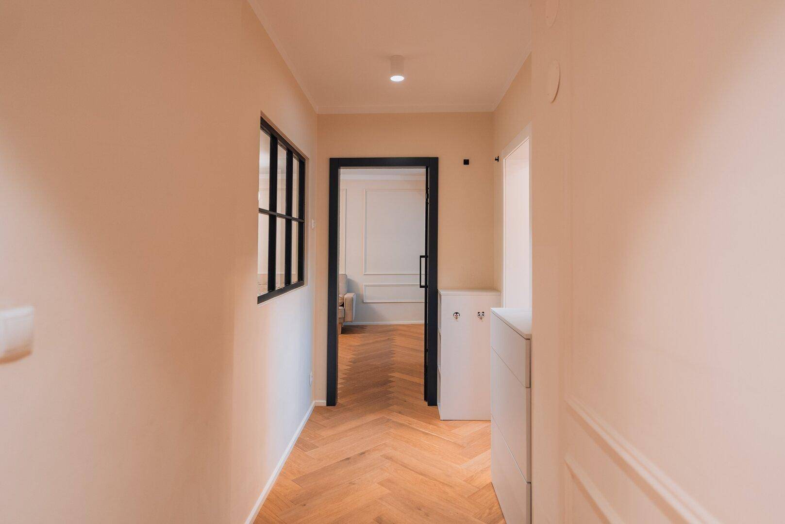 Heller Flur mit Fischgrätparkett, beigen Wänden und einer modernen schwarzen Glastür.