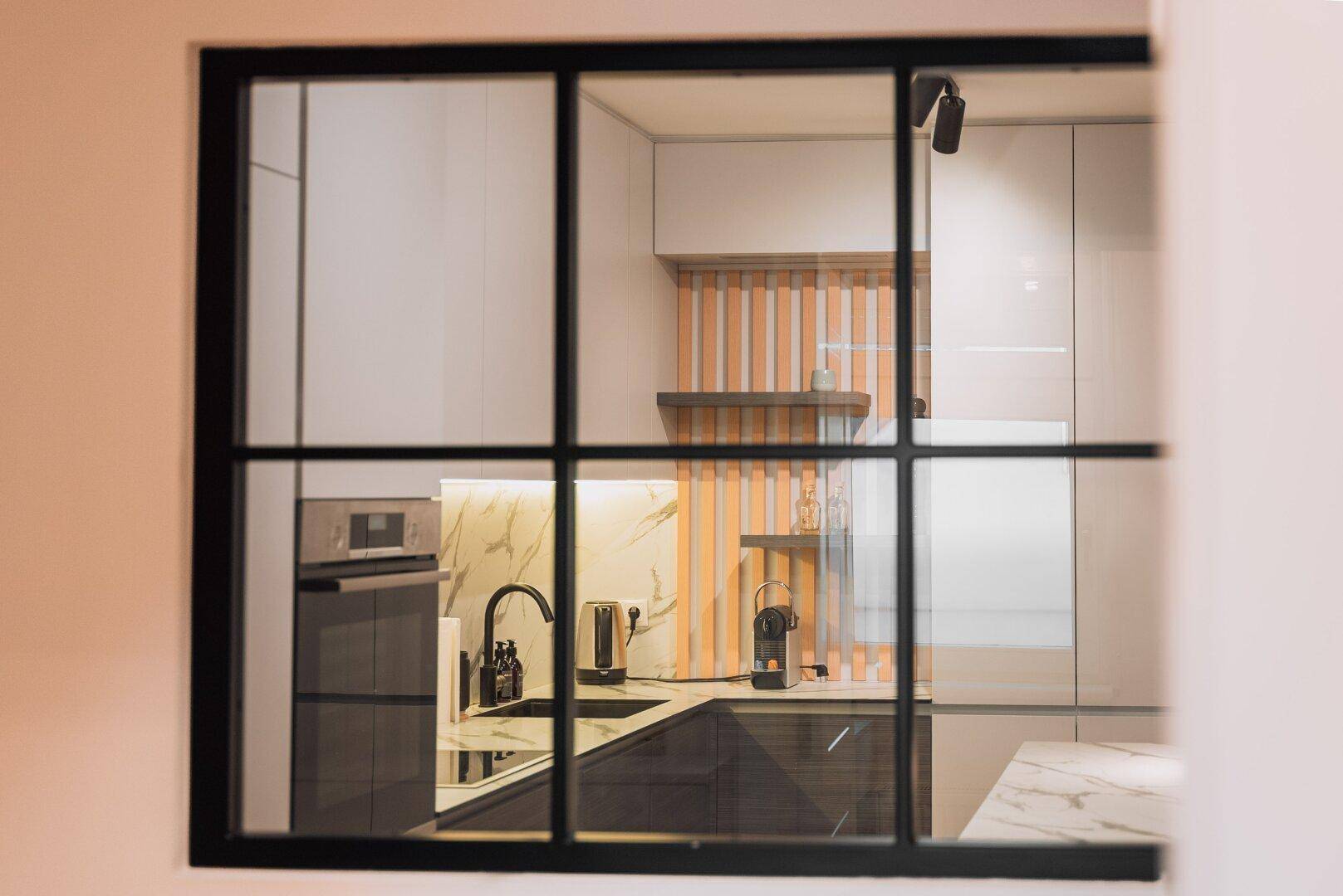 Blick durch ein modernes Fenster in die stilvolle Küche mit Kücheninsel und Marmor-Rückwand.