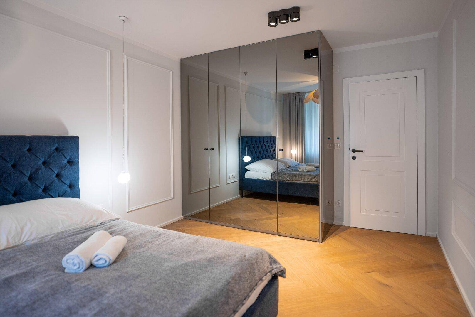 Schlafzimmer mit großem Spiegelschrank, Doppelbett und Fischgrätparkett, das den Raum optisch vergrößert.