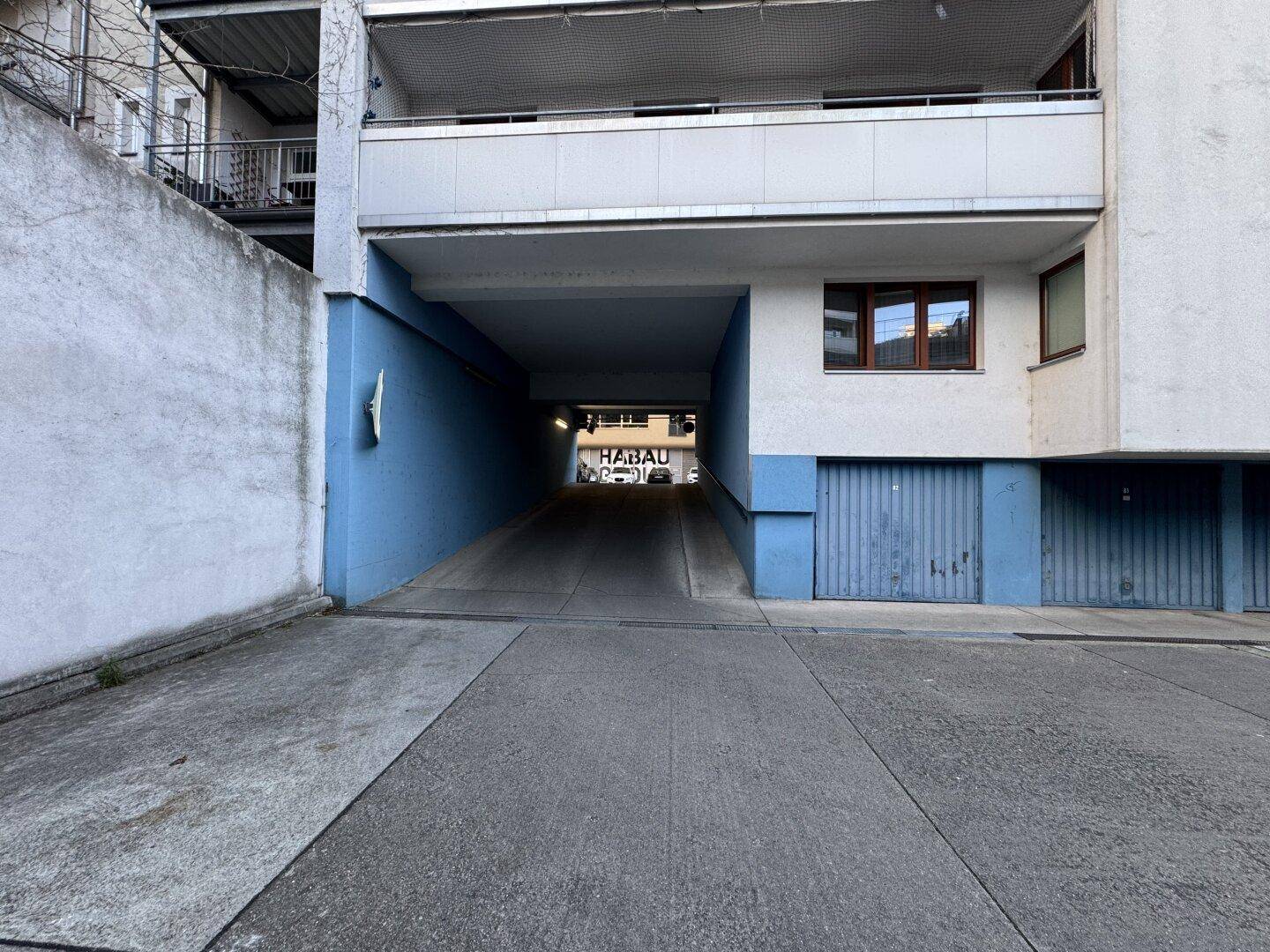 Zufahrt zu einer Tiefgarage oder überdachten Parkfläche mit blauer Fassade des Wohngebäudes.